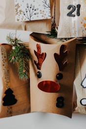 A collection of brown paper gift bags decorated with festive designs, including a reindeer face with red antlers and nose, and numbers like 8 and 21. Sprigs of evergreen are tucked into the bags for decoration. One bag features a black silhouette of a Christmas tree.