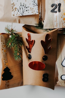 A collection of brown paper gift bags decorated with festive designs, including a reindeer face with red antlers and nose, and numbers like 8 and 21. Sprigs of evergreen are tucked into the bags for decoration. One bag features a black silhouette of a Christmas tree.