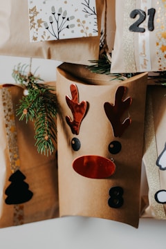 A collection of brown paper gift bags decorated with festive designs, including a reindeer face with red antlers and nose, and numbers like 8 and 21. Sprigs of evergreen are tucked into the bags for decoration. One bag features a black silhouette of a Christmas tree.