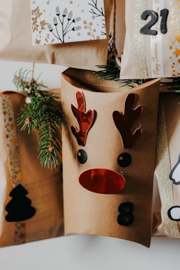A collection of brown paper gift bags decorated with festive designs, including a reindeer face with red antlers and nose, and numbers like 8 and 21. Sprigs of evergreen are tucked into the bags for decoration. One bag features a black silhouette of a Christmas tree.