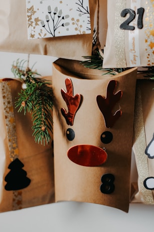 A collection of brown paper gift bags decorated with festive designs, including a reindeer face with red antlers and nose, and numbers like 8 and 21. Sprigs of evergreen are tucked into the bags for decoration. One bag features a black silhouette of a Christmas tree.