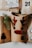 A collection of brown paper gift bags decorated with festive designs, including a reindeer face with red antlers and nose, and numbers like 8 and 21. Sprigs of evergreen are tucked into the bags for decoration. One bag features a black silhouette of a Christmas tree.
