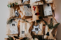 An arrangement of brown paper bags decorated with holiday-themed designs, including stars, bells, and reindeer antlers. Each bag is numbered, possibly for an advent calendar, with small evergreen branches used as additional decoration. The numbers are arranged randomly, suggesting a countdown to Christmas.