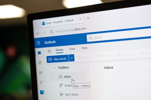 A close-up view of a computer screen displaying the Outlook email client. The image shows the Outlook interface with a tab labeled 'Email - Unsplash - Outlook' and the URL 'https://outlook.office.com'. The interface includes options like 'New email', 'Home', 'View', 'Help', and folder labels such as 'Inbox' and 'Drafts'. The cursor is pointing to the 'Inbox' which shows '0 items'.
