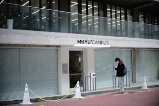 A modern building entrance labeled 'MKYU Campus' features large glass windows and a balcony. A person stands outside, wearing a mask, dark jacket, and backpack, looking at their phone in a relaxed manner.