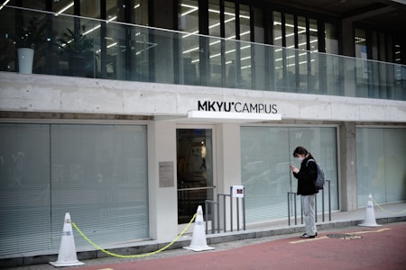 A modern building entrance labeled 'MKYU Campus' features large glass windows and a balcony. A person stands outside, wearing a mask, dark jacket, and backpack, looking at their phone in a relaxed manner.