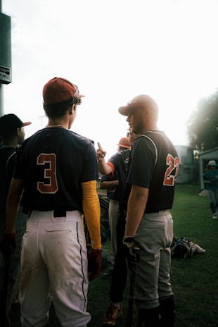 A group of baseball players stand together on a grassy field, wearing uniforms with numbers and caps. They appear to be engaged in conversation or preparing for a game. The lighting casts shadows and creates a bright background, suggesting afternoon or early evening.