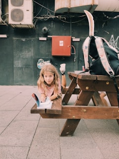 A young child with long hair is lying on a wooden picnic bench outdoors, looking at an open book. A backpack with geometric patterns is placed on the bench beside the child. The setting appears to be urban, with industrial elements like a red fire safety box and air conditioning units mounted on a dark wall in the background.