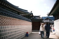A traditional Korean architectural scene with curved roofs and intricate details. A couple walks along a path, surrounded by brick and stone walls. Several people in the distance wear what appears to be traditional Korean attire.
