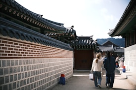 A traditional Korean architectural scene with curved roofs and intricate details. A couple walks along a path, surrounded by brick and stone walls. Several people in the distance wear what appears to be traditional Korean attire.