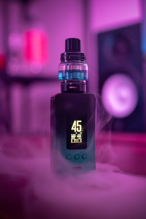 A vape device with a digital display showing the number 45 is surrounded by colorful smoke. The scene is illuminated with purple and blue lighting, giving it a modern and stylish aesthetic. The vape sits on a surface with a blurred background, emphasizing its sleek design.