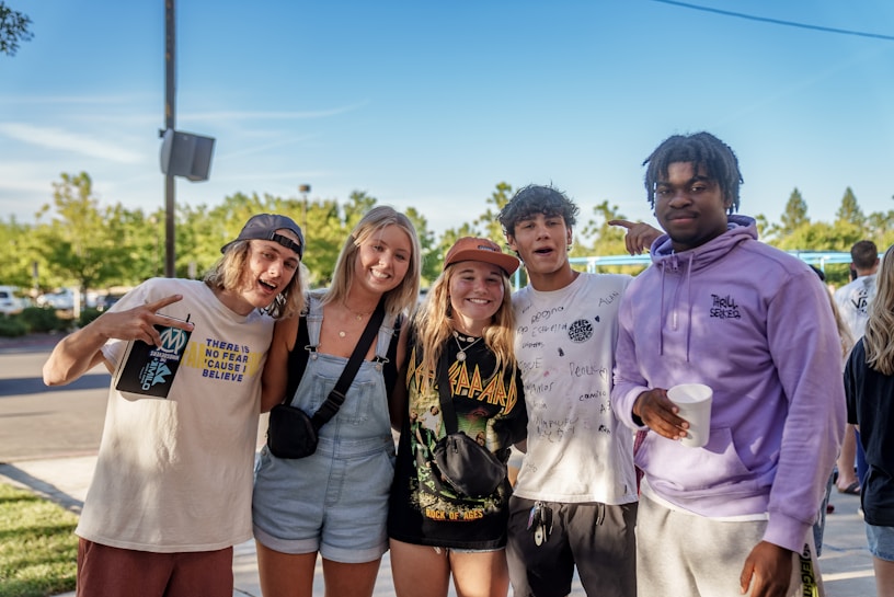 A group of five young adults are standing outdoors, posing for a casual photo. They appear happy and relaxed, with smiles and playful gestures. The setting is a sunny day with a clear blue sky and green trees in the background, suggesting a park or an outdoor event.