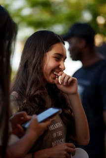 A young woman with long hair is smiling and appearing playful, surrounded by blurred figures in the background. Her hand is near her mouth, and she is wearing a shirt that has text on it. Another person nearby is using a smartphone.