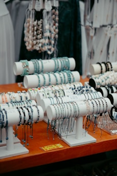 A display of multiple bracelets arranged on stands, featuring various colors and designs. The setup is organized on a wooden table with signs indicating pricing. In the background, there are hanging necklaces.
