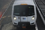 A white train with headlights on, traveling on a railway track. The train has a logo and is flanked by grey gravel and orange construction cones.