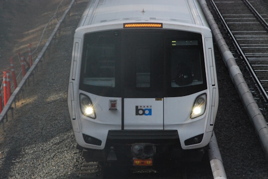 A white train with headlights on, traveling on a railway track. The train has a logo and is flanked by grey gravel and orange construction cones.