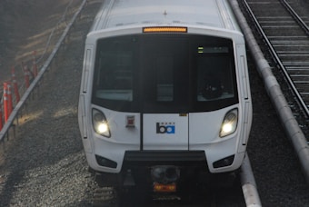 A white train with headlights on, traveling on a railway track. The train has a logo and is flanked by grey gravel and orange construction cones.