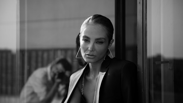 A person stands near a glass window, wearing a stylish blazer. The setting appears urban and sophisticated, with reflections visible in the glass. The monochrome palette adds a classic and timeless feel.