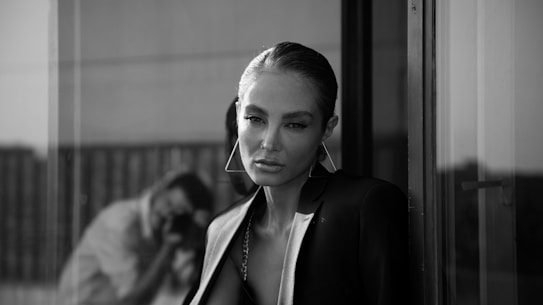 A person stands near a glass window, wearing a stylish blazer. The setting appears urban and sophisticated, with reflections visible in the glass. The monochrome palette adds a classic and timeless feel.