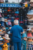 A person is browsing a colorful assortment of hats at an outdoor stand. The hats feature various sports team logos and come in different styles, such as baseball caps and straw hats. The display is vibrant and eye-catching, with multiple shelves filled with neatly arranged headgear.
