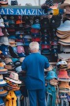A person is browsing a colorful assortment of hats at an outdoor stand. The hats feature various sports team logos and come in different styles, such as baseball caps and straw hats. The display is vibrant and eye-catching, with multiple shelves filled with neatly arranged headgear.