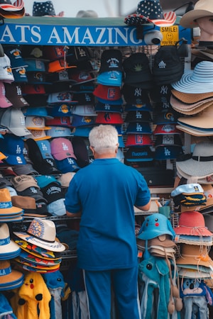 A person is browsing a colorful assortment of hats at an outdoor stand. The hats feature various sports team logos and come in different styles, such as baseball caps and straw hats. The display is vibrant and eye-catching, with multiple shelves filled with neatly arranged headgear.