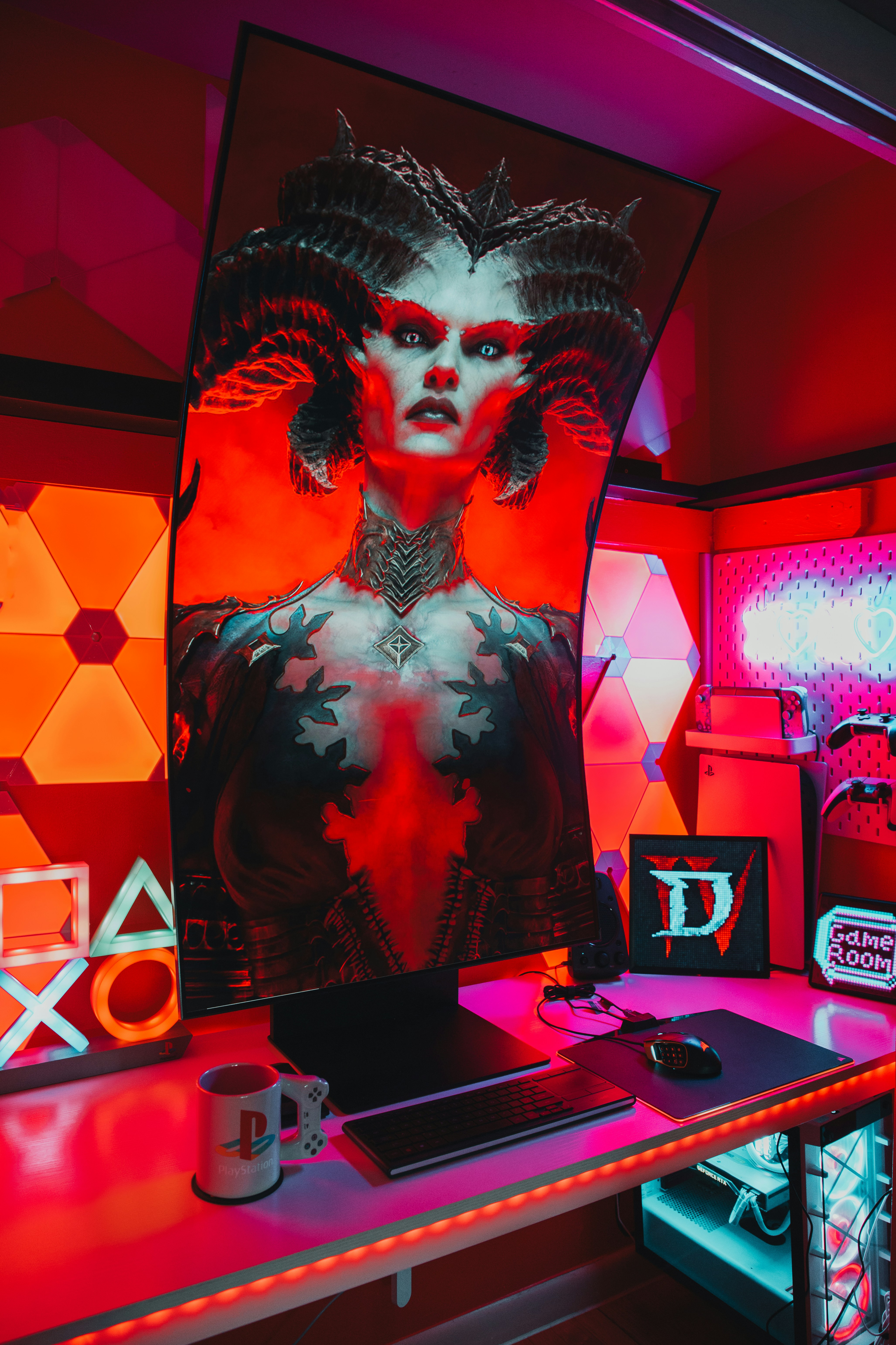 A gaming setup features a large, curved monitor displaying a detailed, fantasy-themed character with horns and vibrant red and black colors. The desk is illuminated with red and purple LED lights, and there are gaming accessories like a keyboard, mouse, and console-themed mug. The background has geometric wall panels and a sign that reads 'Game Room'.