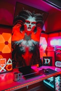 A gaming setup features a large, curved monitor displaying a detailed, fantasy-themed character with horns and vibrant red and black colors. The desk is illuminated with red and purple LED lights, and there are gaming accessories like a keyboard, mouse, and console-themed mug. The background has geometric wall panels and a sign that reads 'Game Room'.
