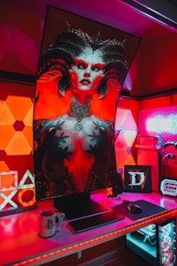 A gaming setup features a large, curved monitor displaying a detailed, fantasy-themed character with horns and vibrant red and black colors. The desk is illuminated with red and purple LED lights, and there are gaming accessories like a keyboard, mouse, and console-themed mug. The background has geometric wall panels and a sign that reads 'Game Room'.