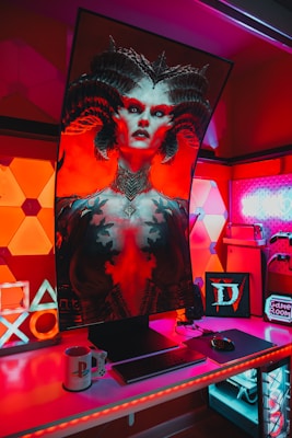 A gaming setup features a large, curved monitor displaying a detailed, fantasy-themed character with horns and vibrant red and black colors. The desk is illuminated with red and purple LED lights, and there are gaming accessories like a keyboard, mouse, and console-themed mug. The background has geometric wall panels and a sign that reads 'Game Room'.