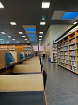A spacious library interior with shelves filled with books along the walls. There are several wooden study cubicles with blue reading lamps attached to them. The library has a high ceiling with bright rectangular lights and blue skylights that allow natural light to enter. The floor is a gray, polished surface, contributing to the modern and clean aesthetic.