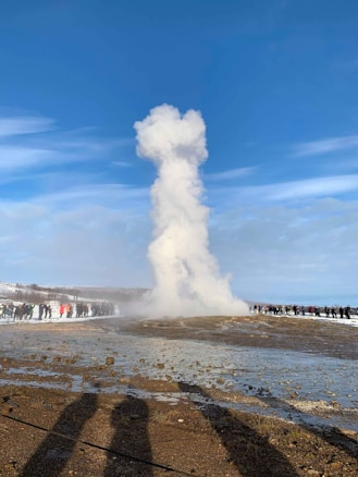 A large geyser erupts, spewing a thick column of steam and water into the clear blue sky. A crowd of people stand at a distance, observing the natural phenomenon on a wet, rocky surface surrounded by snow. The scene is bathed in sunlight, casting long shadows on the ground.