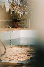 An outdoor swimming pool that is mostly empty, with a small amount of water and scattered leaves on the bottom. The pool is surrounded by a fence and there are trees in the background with autumn foliage. The sunlight casts a warm glow, and the scene is partially obscured, giving it a dreamy and nostalgic feel.