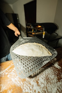 A person is using a metal pizza peel to handle a piece of dough covered in flour, with a wood-fired oven in the background. The surface is dusted with flour, indicating preparation for baking.