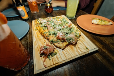 A flatbread with toppings such as prosciutto, greens, and grated cheese is served on a wooden board. Nearby, a partially eaten breadstick lies on an orange plate. Various colored drinks in glasses and bottles are arranged across the dark wooden table, along with blue plates and a small jar.