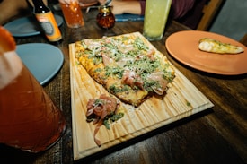 A flatbread with toppings such as prosciutto, greens, and grated cheese is served on a wooden board. Nearby, a partially eaten breadstick lies on an orange plate. Various colored drinks in glasses and bottles are arranged across the dark wooden table, along with blue plates and a small jar.