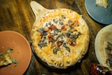 A freshly baked pizza topped with mushrooms, cheese, and vegetables is placed on a wooden serving paddle. The surrounding area features a rustic wooden table setting with two plates nearby, one partially eaten slice of pizza on a plate, and a bottle of chili flakes.