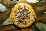 A freshly baked pizza topped with a generous amount of roasted vegetables, including mushrooms and red peppers, rests on a rustic wooden table. The pizza has a golden-brown crust and is served on a wooden paddle. The table setting includes a variety of glassware and dishes, hinting at a cozy dining atmosphere.