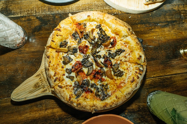 A freshly baked pizza topped with a generous amount of roasted vegetables, including mushrooms and red peppers, rests on a rustic wooden table. The pizza has a golden-brown crust and is served on a wooden paddle. The table setting includes a variety of glassware and dishes, hinting at a cozy dining atmosphere.