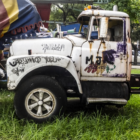 A rusty old truck is parked on grassy ground, with a worn-out exterior covered in graffiti and peeling paint. The side of the truck features various graffiti tags and patches of rust. The back of the truck appears to be loaded with assorted items covered by a colorful tarp.