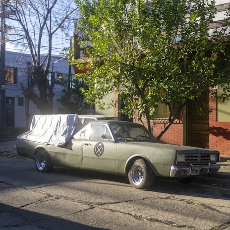 A vintage green car is parked on the side of a suburban street, partially covered with a white tarp. A tree with lush green leaves shades part of the car, while the background includes a residential building with brick and plaster exterior walls. Bare tree branches are visible against a clear blue sky.