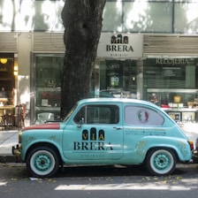 A vintage light blue car with branding parked on the street in front of a jewelry store. The vehicle bears the 'Via Brera' logo and has a retro design, complementing the urban setting. Trees cast shadows on the sidewalk, and the store windows display various items.
