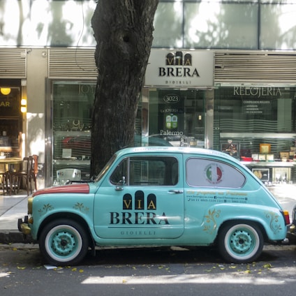 A vintage light blue car with branding parked on the street in front of a jewelry store. The vehicle bears the 'Via Brera' logo and has a retro design, complementing the urban setting. Trees cast shadows on the sidewalk, and the store windows display various items.