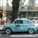 A vintage light blue car with branding parked on the street in front of a jewelry store. The vehicle bears the 'Via Brera' logo and has a retro design, complementing the urban setting. Trees cast shadows on the sidewalk, and the store windows display various items.