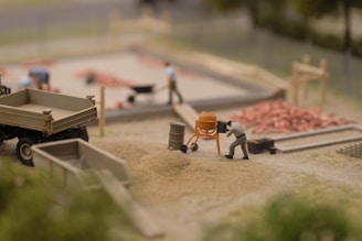 A miniature construction scene shows a person operating a small cement mixer near piles of sand and gravel. There are other figures in the background, some working with a wheelbarrow and others around a construction area with visible piles of red bricks. A miniature truck and various construction tools are also present.