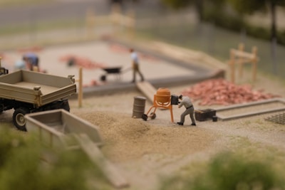 A miniature construction scene shows a person operating a small cement mixer near piles of sand and gravel. There are other figures in the background, some working with a wheelbarrow and others around a construction area with visible piles of red bricks. A miniature truck and various construction tools are also present.