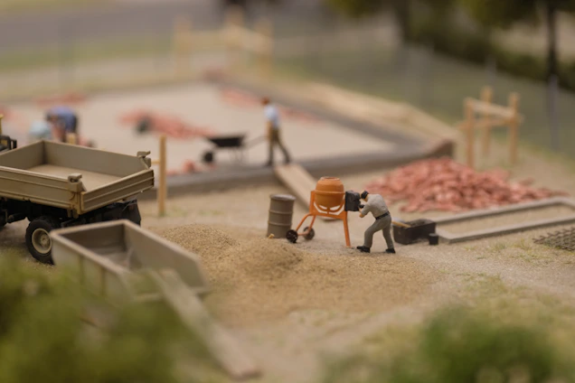 A miniature construction scene shows a person operating a small cement mixer near piles of sand and gravel. There are other figures in the background, some working with a wheelbarrow and others around a construction area with visible piles of red bricks. A miniature truck and various construction tools are also present.