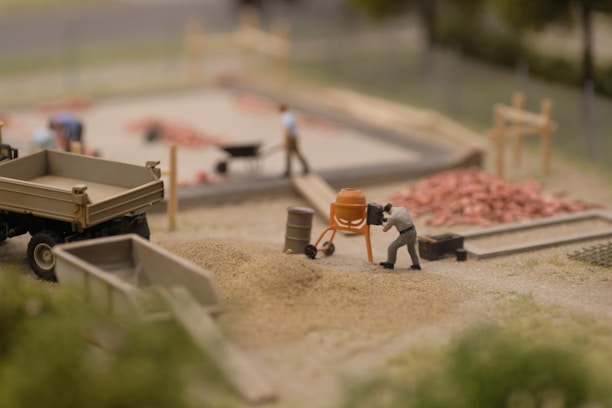 A miniature construction scene shows a person operating a small cement mixer near piles of sand and gravel. There are other figures in the background, some working with a wheelbarrow and others around a construction area with visible piles of red bricks. A miniature truck and various construction tools are also present.