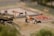 A miniature construction scene shows a person operating a small cement mixer near piles of sand and gravel. There are other figures in the background, some working with a wheelbarrow and others around a construction area with visible piles of red bricks. A miniature truck and various construction tools are also present.