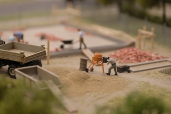 A miniature construction scene shows a person operating a small cement mixer near piles of sand and gravel. There are other figures in the background, some working with a wheelbarrow and others around a construction area with visible piles of red bricks. A miniature truck and various construction tools are also present.